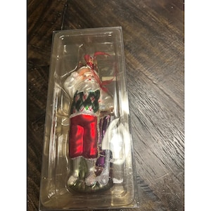 May include: A Christmas ornament of Santa Claus dressed in a golf outfit. The ornament features a white beard and hair, a red and green argyle sweater vest, red pants, and a golf club. The ornament is in a clear plastic case.