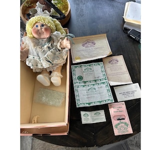 May include: A Cabbage Patch Kids doll with blonde yarn hair, blue eyes, and a white dress with blue accents. The doll is in a pink box with several certificates and papers.