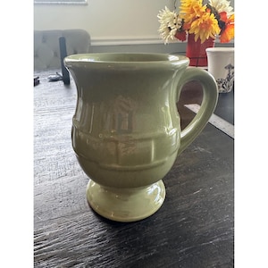 Puede incluir: Una taza de cerámica color verde salvia con acabado brillante, que presenta un borde ancho y acampanado, un cuerpo redondeado con un sutil patrón de rejilla en relieve, un asa lateral resistente y una base de pedestal. Superficie lisa y brillante.