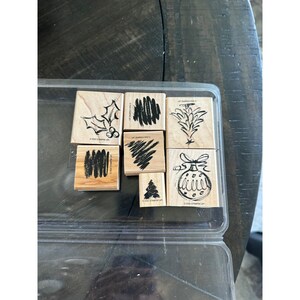 Op de afbeelding: Een verzameling houten stempels met diverse vakantie-thema ontwerpen, waaronder hulst, abstracte patronen en een kerstbal. De stempels zijn gerangschikt in een doorzichtige plastic container.