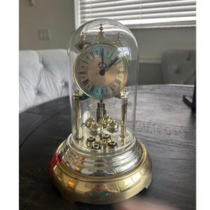 Puede incluir: Un reloj de aniversario dorado con una esfera circular con números romanos y manecillas negras, encerrado en una cúpula de cristal transparente. El reloj descansa sobre una base dorada pulida e incluye un péndulo giratorio con cuatro esferas.