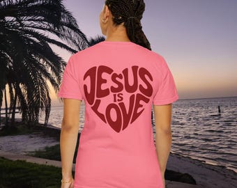 May include: A pink t-shirt with the words "JESUS IS LOVE" in a heart shape on the back. The text is a deep red color. The shirt is being worn in front of a beach scene with palm trees and a sunset.