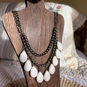 May include: A multi-strand necklace with a bronze-colored chain and teardrop-shaped, off-white stone pendants. The necklace is displayed on a wooden jewelry stand, showcasing its layered design and textured details.