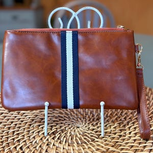 May include: A brown leather clutch with a navy and white stripe down the center. The clutch has a wrist strap and a gold zipper. The clutch is displayed on a woven wicker surface.