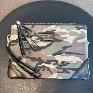 May include: Camouflage print clutch with black leather accents. The rectangular bag features a zipper closure, a wrist strap, and a black leather label that reads "FUNKY MONKEY". The bag is in shades of green, brown, and tan.