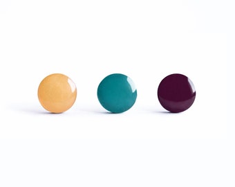 May include: Three glossy, round objects in a row against a white background. The objects are in shades of yellow, teal, and deep purple. The objects appear to be nail polish or similar beauty products.