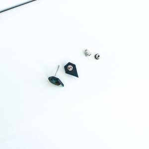 May include: A pair of stud earrings with a geometric design. One earring features a faceted, iridescent gem, while the other has a dark blue, shield-shaped backing. The earrings are accompanied by two silver earring backs, all set against a white background.