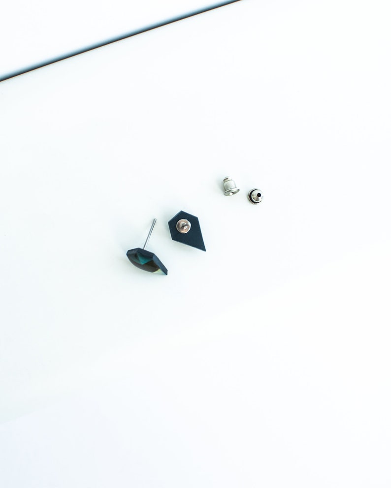 May include: A pair of stud earrings with a geometric design. One earring features a faceted, iridescent gem, while the other has a dark blue, shield-shaped backing. The earrings are accompanied by two silver earring backs, all set against a white background.