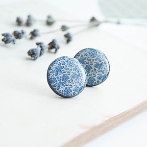 Pode incluir: Dois brincos redondos com um padr&atilde;o floral azul em fundo branco. Os brincos est&atilde;o sobre uma superf&iacute;cie de madeira clara, com raminhos de lavanda seca ao fundo.