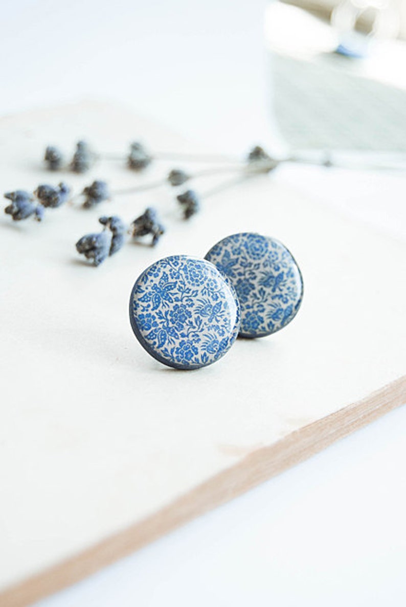 Pode incluir: Dois brincos redondos com um padr&atilde;o floral azul em fundo branco. Os brincos est&atilde;o sobre uma superf&iacute;cie de madeira clara, com raminhos de lavanda seca ao fundo.