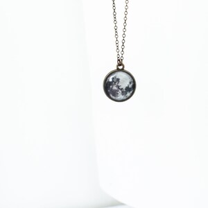 May include: A pendant necklace featuring a round charm with a detailed image of the moon. The charm is suspended from a delicate, antique bronze-colored chain. The moon's surface is depicted in shades of gray and white.