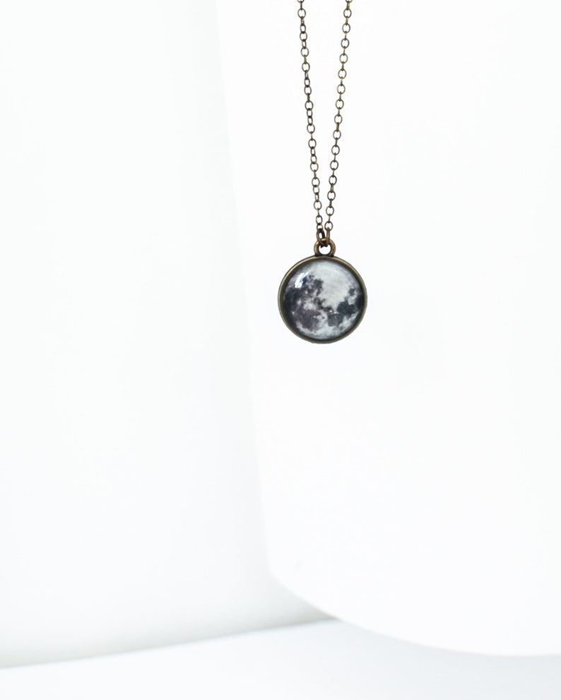 May include: A pendant necklace featuring a round charm with a detailed image of the moon. The charm is suspended from a delicate, antique bronze-colored chain. The moon's surface is depicted in shades of gray and white.