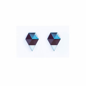 May include: A pair of geometric stud earrings. Each earring features a dark burgundy, teal, and white design. The earrings have a diamond shape with a pointed bottom. The background is white.