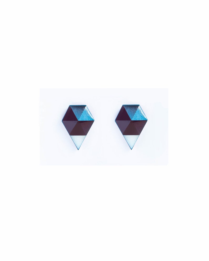 May include: A pair of geometric stud earrings. Each earring features a dark burgundy, teal, and white design. The earrings have a diamond shape with a pointed bottom. The background is white.