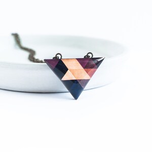 May include: Geometric triangle pendant necklace. The pendant features a mosaic design with shades of purple, navy, and peach. The necklace has a dark chain and is set against a white background.
