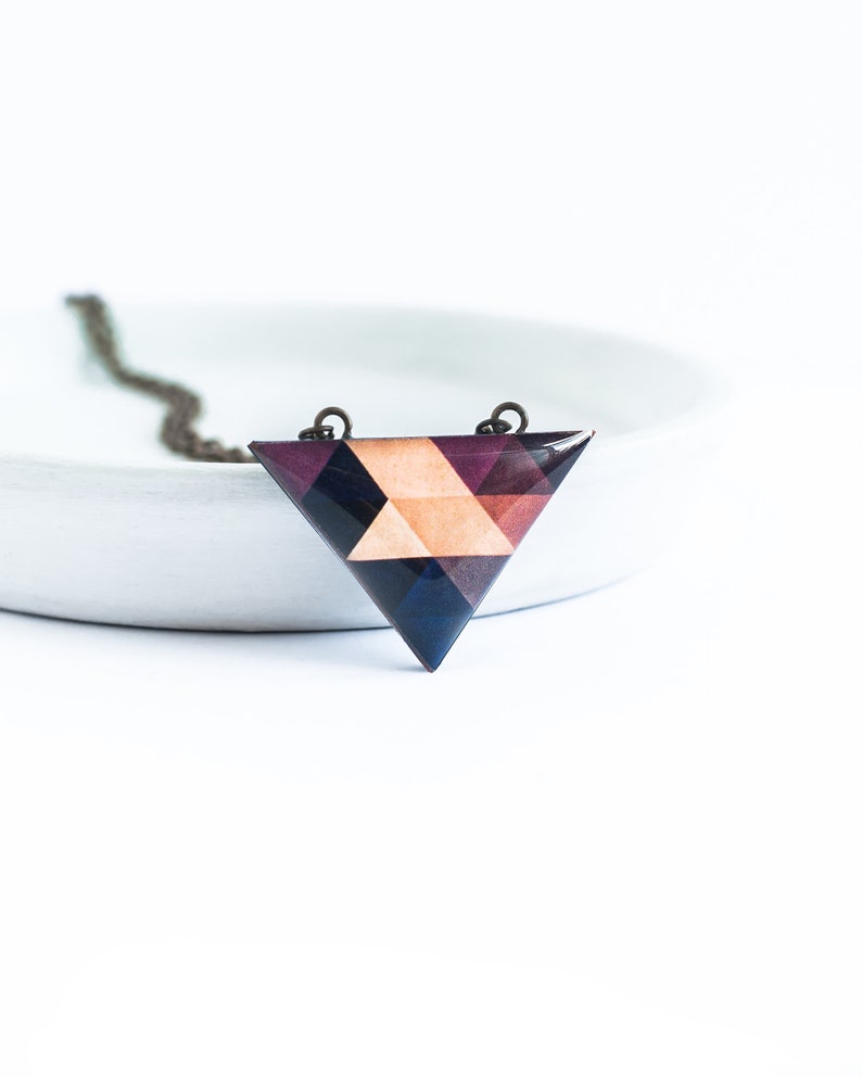 May include: Geometric triangle pendant necklace. The pendant features a mosaic design with shades of purple, navy, and peach. The necklace has a dark chain and is set against a white background.