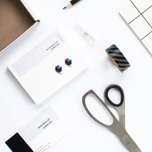 May include: A flat lay featuring minimalist jewelry, stationery, and craft supplies. The focal point is a pair of geometric stud earrings on a white card. Other items include a pencil, scissors, washi tape, and a notepad, all arranged on a white surface.
