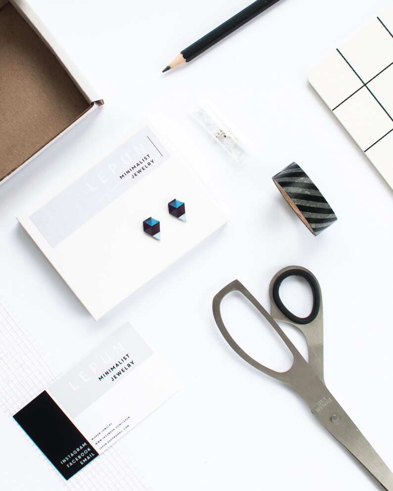 May include: A flat lay featuring minimalist jewelry, stationery, and craft supplies. The focal point is a pair of geometric stud earrings on a white card. Other items include a pencil, scissors, washi tape, and a notepad, all arranged on a white surface.
