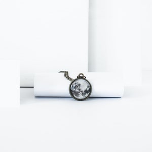 May include: A round pendant necklace featuring a detailed grayscale image of the moon. The pendant is set in a bronze-colored metal frame and hangs from a matching chain. The necklace is displayed on a white background.