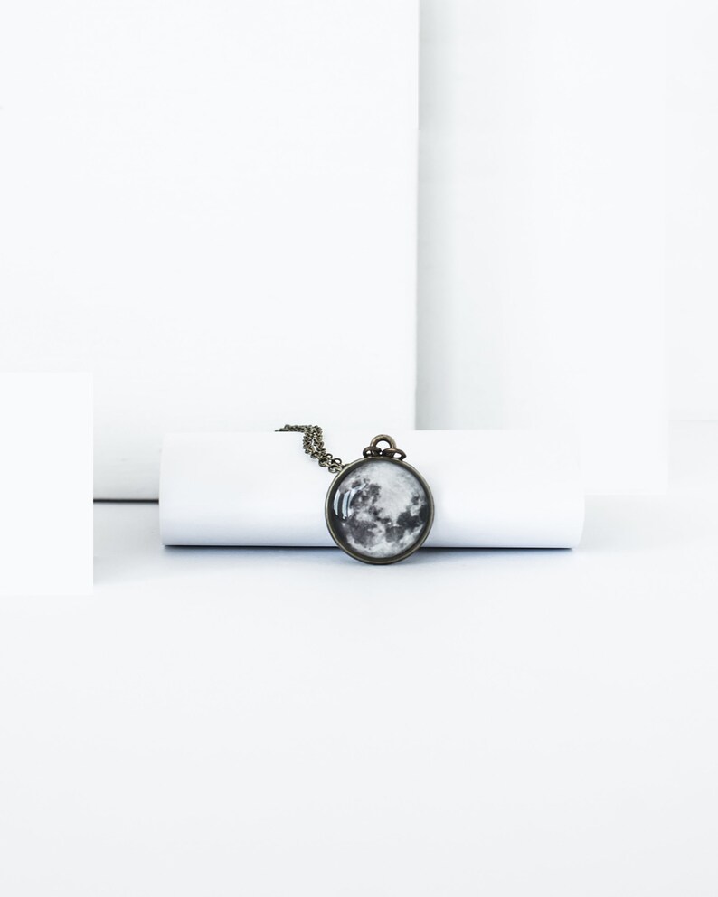 May include: A round pendant necklace featuring a detailed grayscale image of the moon. The pendant is set in a bronze-colored metal frame and hangs from a matching chain. The necklace is displayed on a white background.