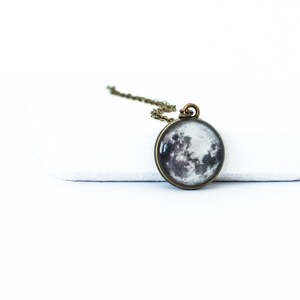 May include: A round pendant necklace featuring a detailed image of the moon. The pendant is set in a bronze-colored metal frame and hangs from a matching chain. The moon image displays shades of gray and white, creating a realistic lunar surface.