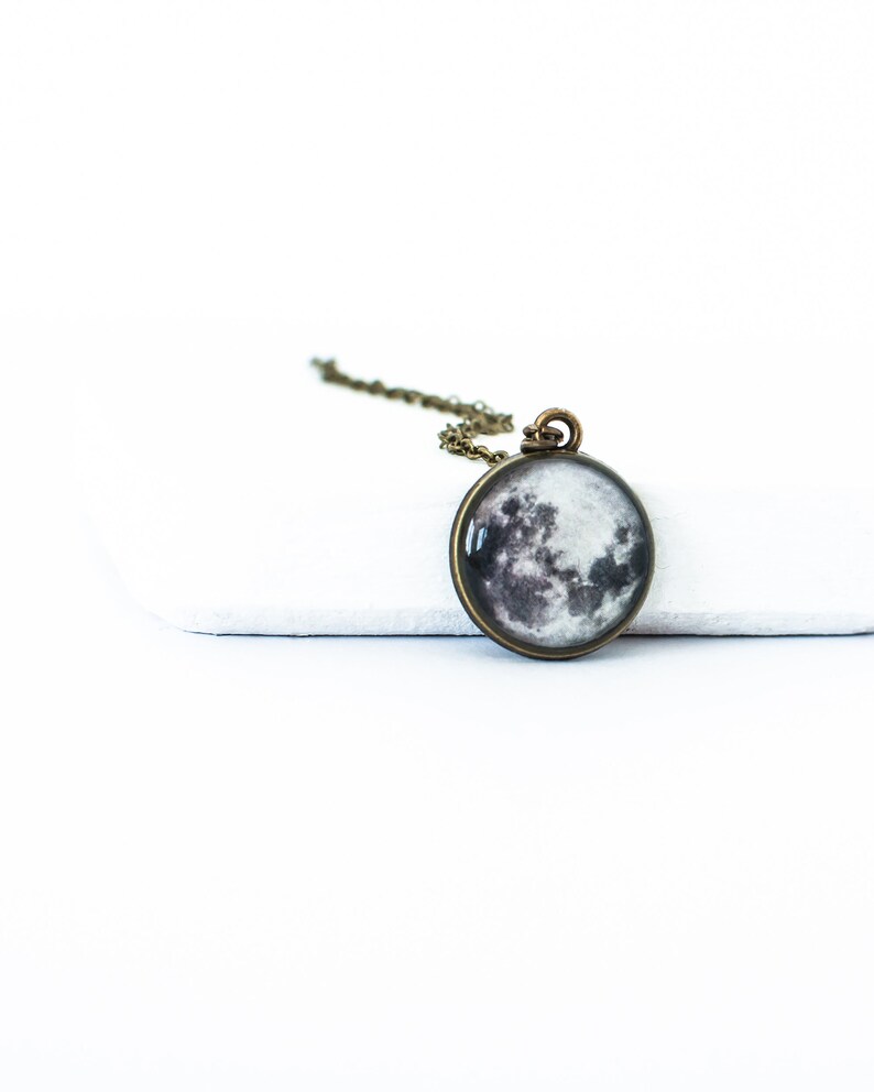 May include: A round pendant necklace featuring a detailed image of the moon. The pendant is set in a bronze-colored metal frame and hangs from a matching chain. The moon image displays shades of gray and white, creating a realistic lunar surface.
