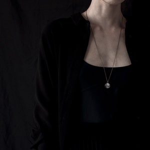 May include: A long, delicate necklace with a circular pendant hangs against a dark background. The pendant is silver-toned and appears to have a detailed design. The person is wearing a black cardigan over a black top.