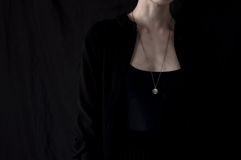 May include: A long, delicate necklace with a circular pendant hangs against a dark background. The pendant is silver-toned and appears to have a detailed design. The person is wearing a black cardigan over a black top.