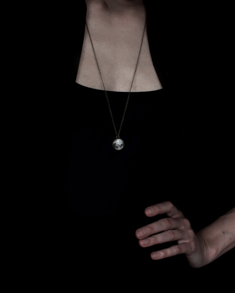May include: A silver pendant necklace with a round charm depicting a detailed moon. The necklace has a delicate chain and is displayed against a black background. The charm is approximately 0.75 inches in diameter.