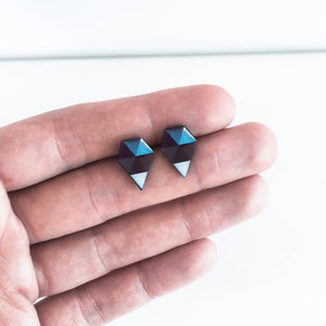 May include: A pair of geometric stud earrings. Each earring is a dark blue and purple diamond shape with a white triangle at the bottom. The earrings are displayed on a light background.
