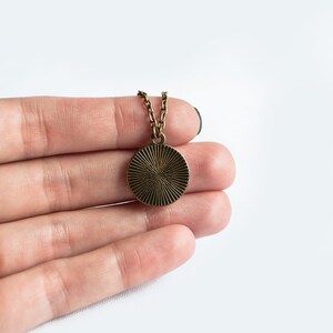 May include: A bronze-colored pendant necklace with a circular, sunburst design. The pendant is suspended from a delicate chain. The necklace is held in a hand against a white background. Jewelry.