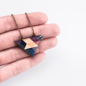 May include: A geometric pendant necklace with a triangular pendant. The pendant features a mosaic design with shades of blue, purple, and tan. The chain is a bronze color. The necklace is held in a hand against a white background.