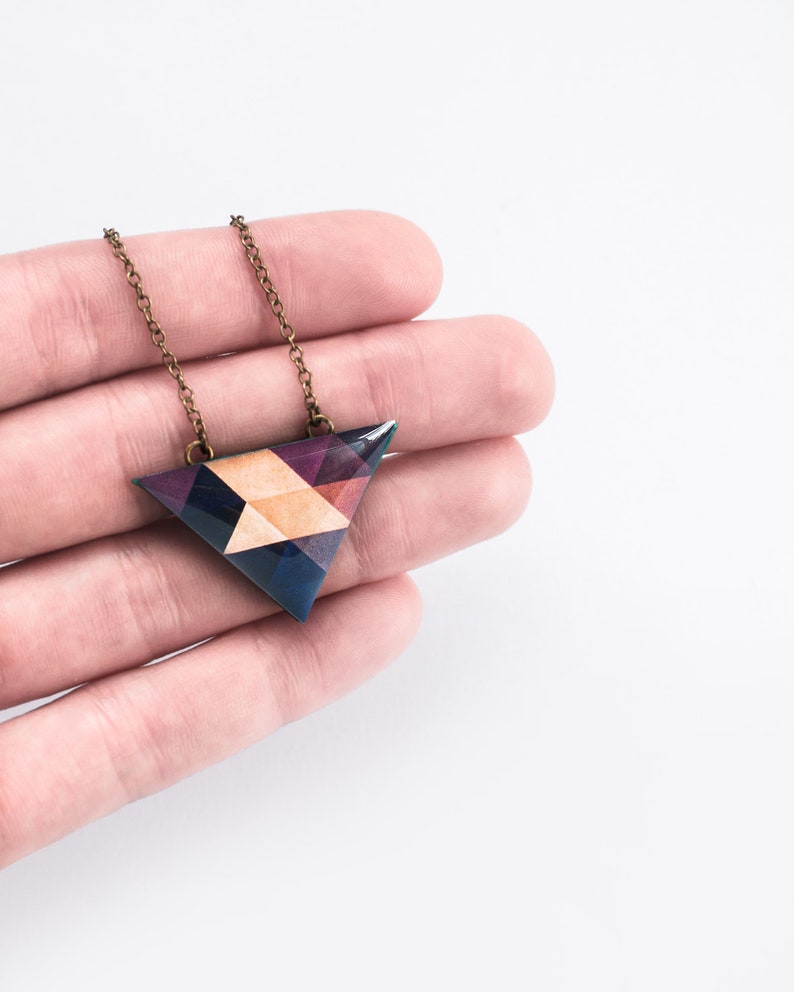 May include: A geometric pendant necklace with a triangular pendant. The pendant features a mosaic design with shades of blue, purple, and tan. The chain is a bronze color. The necklace is held in a hand against a white background.
