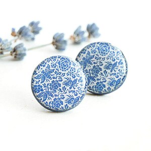 Pode incluir: Dois brincos redondos com um padr&atilde;o floral azul e branco. Os brincos t&ecirc;m um acabamento brilhante e uma borda azul escura. Um raminho de lavanda est&aacute; no fundo.