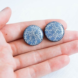 Pode incluir: Dois brincos redondos com um padr&atilde;o floral azul sobre um fundo branco. Os brincos t&ecirc;m uma borda preta e s&atilde;o segurados em uma m&atilde;o. O design floral apresenta flores e folhas.