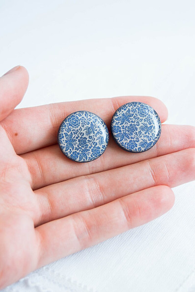 Pode incluir: Dois brincos redondos com um padr&atilde;o floral azul sobre um fundo branco. Os brincos t&ecirc;m uma borda preta e s&atilde;o segurados em uma m&atilde;o. O design floral apresenta flores e folhas.