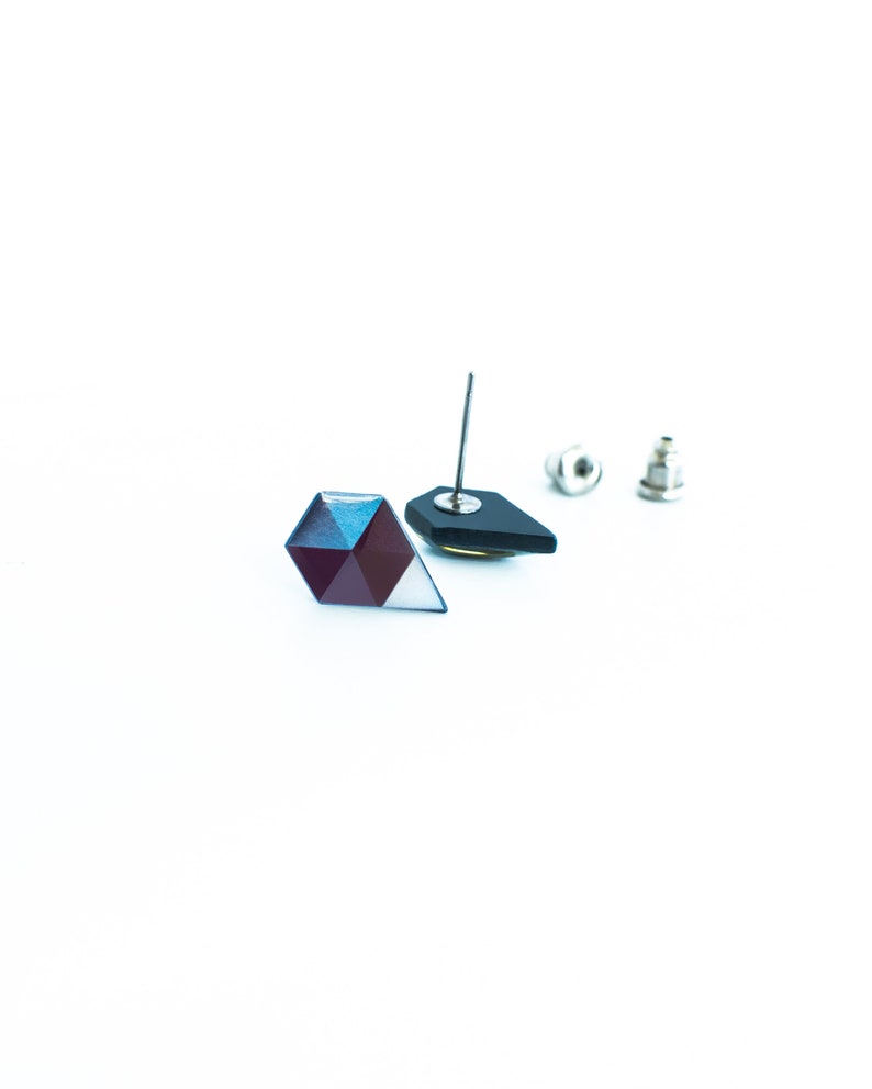 May include: Geometric stud earrings with a faceted, multi-colored design. One earring is a dark, angular shape with a silver post. The other earring is a multi-faceted shape with shades of blue, purple, and white. The earring backs are also visible.