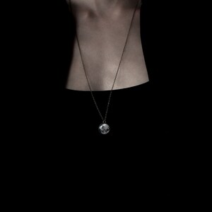 May include: A silver-toned pendant necklace with a round charm. The charm features a detailed, possibly photographic, image. The necklace hangs against a dark background, highlighting the pendant's design.