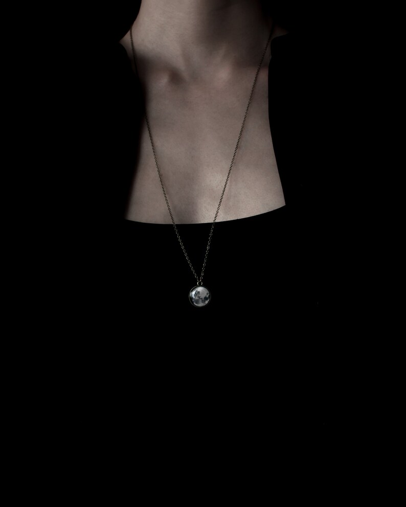 May include: A silver-toned pendant necklace with a round charm. The charm features a detailed, possibly photographic, image. The necklace hangs against a dark background, highlighting the pendant's design.