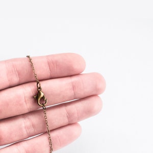May include: Close-up of a bronze-colored necklace chain with a lobster clasp. The chain is held against a white background, showcasing the intricate links and the clasp's design. The chain is a delicate accessory.