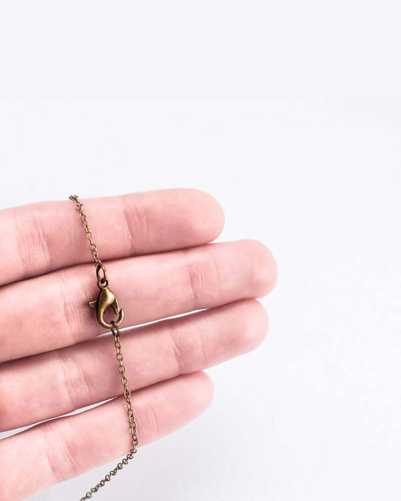 May include: Close-up of a bronze-colored necklace chain with a lobster clasp. The chain is held against a white background, showcasing the intricate links and the clasp's design. The chain is a delicate accessory.