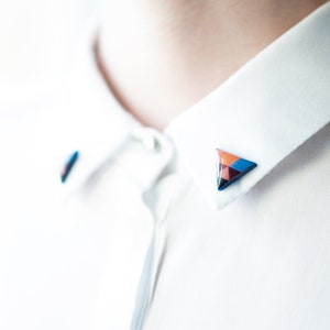 Puede incluir: Primer plano de una camisa blanca con cuello con dos alfileres de cuello triangulares geométricos. Los alfileres presentan un diseño de mosaico con tonos de naranja, azul y burdeos. La camisa está hecha de tela blanca.