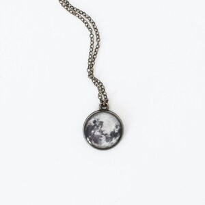 May include: A round pendant necklace featuring a detailed image of the moon. The pendant is set in a silver-colored metal frame and hangs from a delicate chain. The moon image displays shades of gray and white, creating a realistic celestial design.