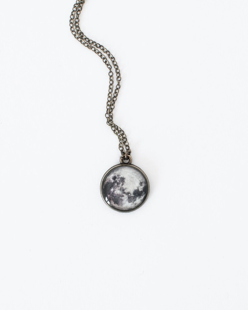 May include: A round pendant necklace featuring a detailed image of the moon. The pendant is set in a silver-colored metal frame and hangs from a delicate chain. The moon image displays shades of gray and white, creating a realistic celestial design.
