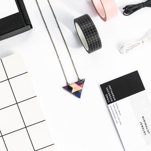 May include: A geometric pendant necklace with a triangular design in shades of purple, blue, and peach. The necklace is on a delicate chain. The image also includes rolls of pink and black washi tape, black and white string, and a business card.