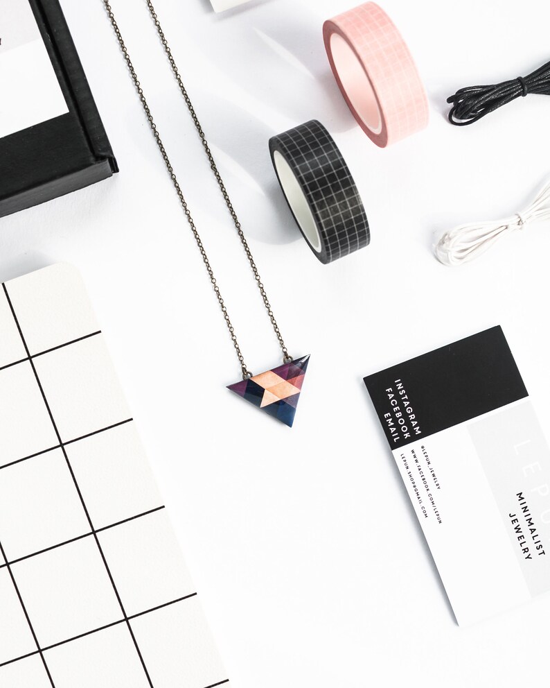May include: A geometric pendant necklace with a triangular design in shades of purple, blue, and peach. The necklace is on a delicate chain. The image also includes rolls of pink and black washi tape, black and white string, and a business card.