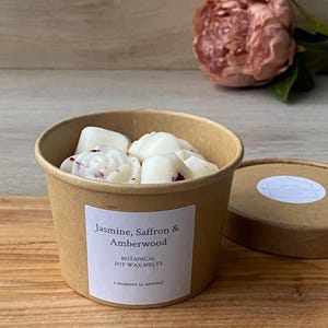 May include: A round, brown container filled with white soy wax melts, some with red accents. The container has a label that reads "Jasmine, Saffron & Amberwood" and "Botanical Soy Wax Melts". A pink flower is in the background.