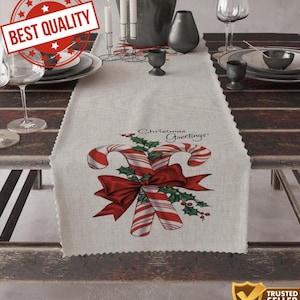 Puede incluir: Camino de mesa navideño color crema con un diseño festivo. Presenta dos bastones de caramelo rojos y blancos, un lazo rojo y hojas de acebo. Las palabras "Christmas Greetings" están impresas encima de los bastones de caramelo. El camino de mesa está sobre una mesa de madera.