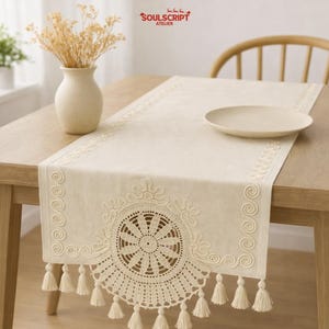 Puede incluir: Camino de mesa color crema con intrincados detalles de encaje y borlas. El camino de mesa presenta un diseño central de encaje circular y bordes decorativos. El logotipo de Soulscript Atelier es visible. Un jarrón con flores secas y un plato están sobre la mesa.