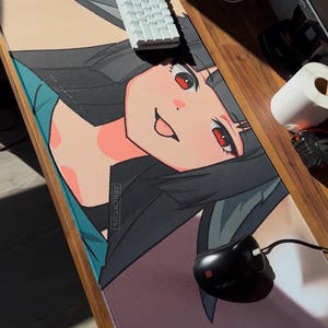 Pode incluir: Um grande tapete de rato retangular com uma ilustração em estilo anime de um personagem sorridente com cabelo preto e olhos vermelhos. Um teclado branco e um rato preto estão no tapete. O tapete tem uma borda azul-turquesa e o texto "2PRCNT.US".